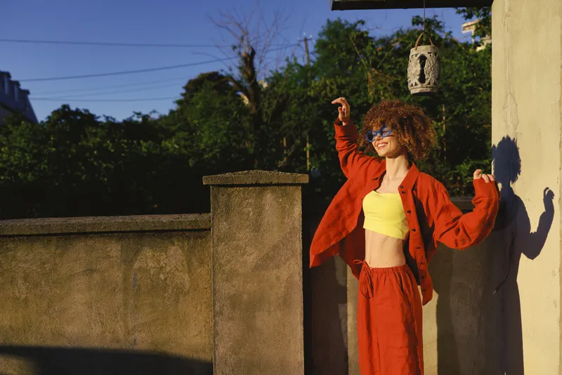 Bright outdoor portrait of a woman in a yellow crop top and red jacket posing confidently on a sunny terrace. Fashion lifestyle concept highlighting vibrant color and casual style
