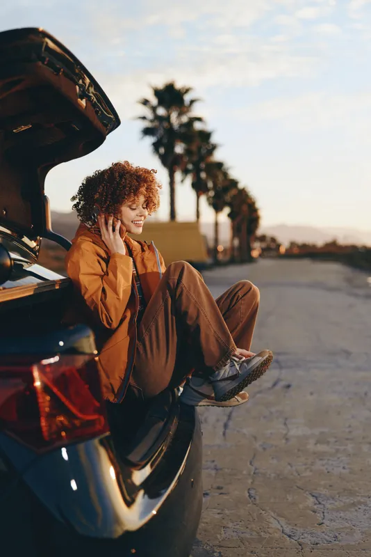 Woman lifestyle outdoors at sunset beside open car, casual jacket and jeans, curly hair, sitting on pavement with relaxed smile, warm golden light, palm lined street background, quiet mood