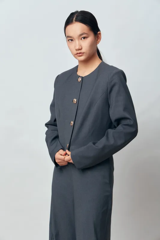 Young woman in gray formal outfit with long sleeves buttons standing against light background looking at camera with serious expression. Studio portrait of female model with natural makeup and