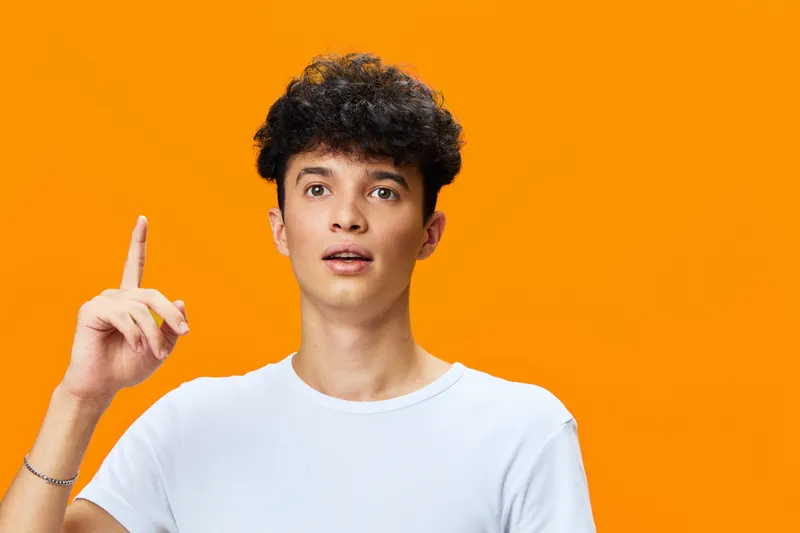 Young man with curly hair wearing white t-shirt pointing up with surprised expression on vibrant orange background, showing creativity and inspiration.