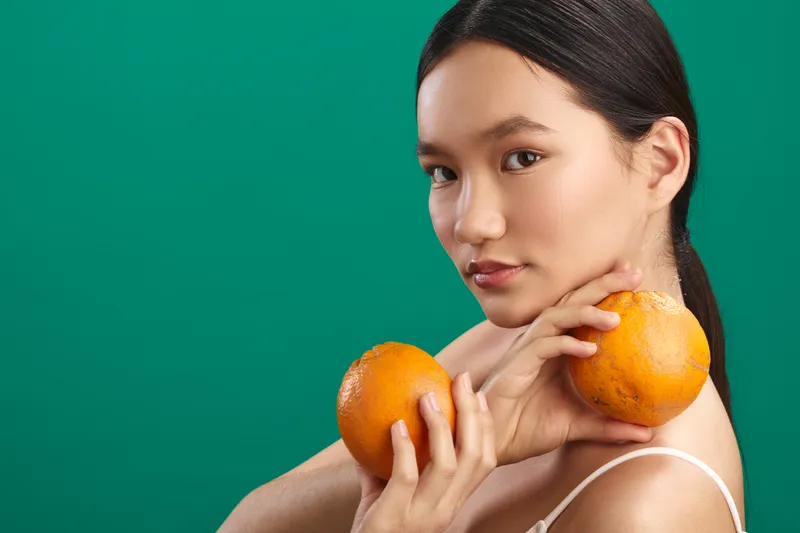 asian woman holds oranges in a studio setting, presenting vibrant fruit and a fresh, healthy lifestyle. the calm expression and bright color create a positive, energetic mood.