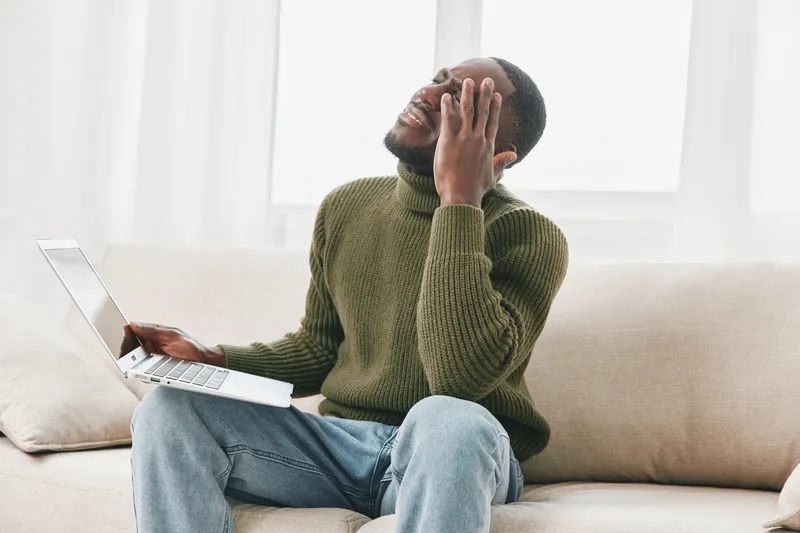 A frustrated young Black man in a green turtleneck sitting on a couch, holding his laptop in one hand and rubbing his forehead, depicting a sense of anxiety and stress