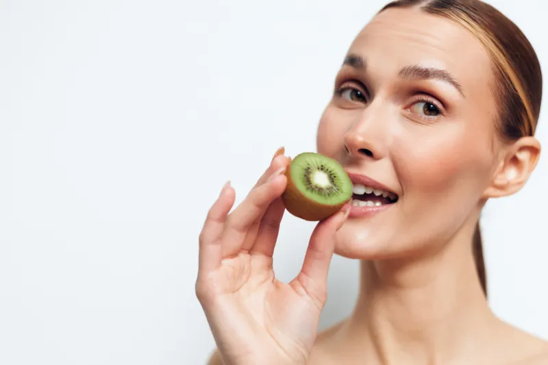Young woman holding kiwi slice near mouth, fresh fruit snack, healthy lifestyle concept, natural skin, bright background, closeup portrait, smiling expression