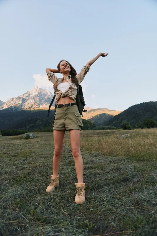 A young woman with long hair wearing a tied shirt and shorts stretches her arms joyfully in a mountain meadow during a clear day, expressing freedom and happiness.