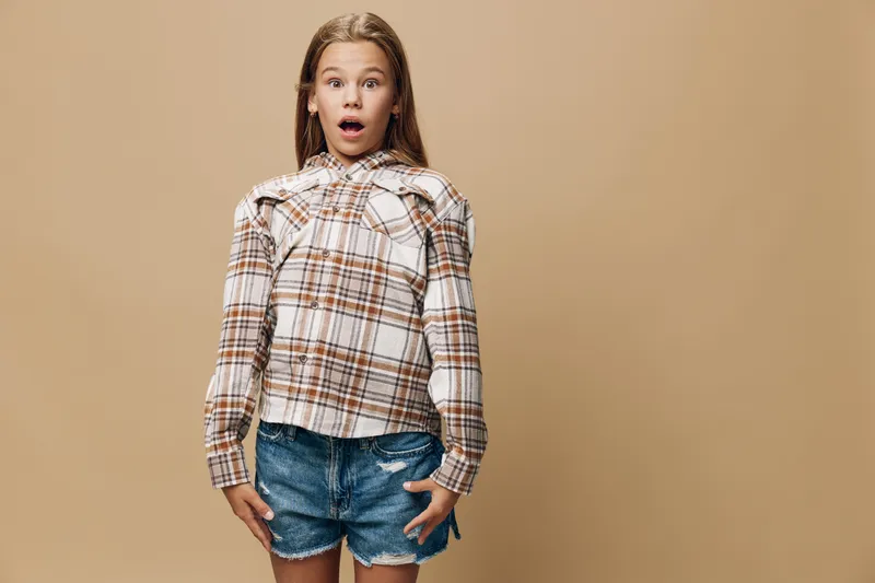Girl with surprised expression standing against beige background wearing plaid shirt and denim shorts, looking shocked with mouth open and wide eyes in casual outfit.
