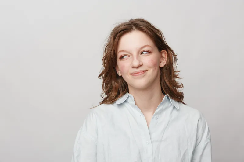 Young woman with wavy brown hair wearing a white shirt looks sideways with a slight smile on a light grey background. Concept of casual style, natural beauty, and positive mood.