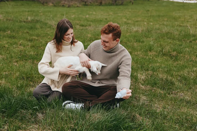 A joyful couple sits on grass in a meadow, holding a goat kid and sharing a warm, gentle moment outdoors. An intimate, rural scene of bonding, happiness, and nature in a peaceful family setting.