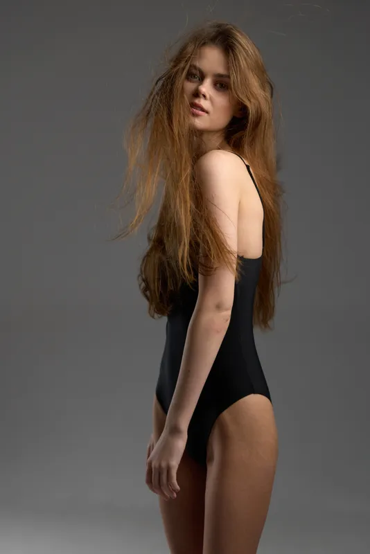 young woman in black swimsuit posing with neutral expression against gray background
