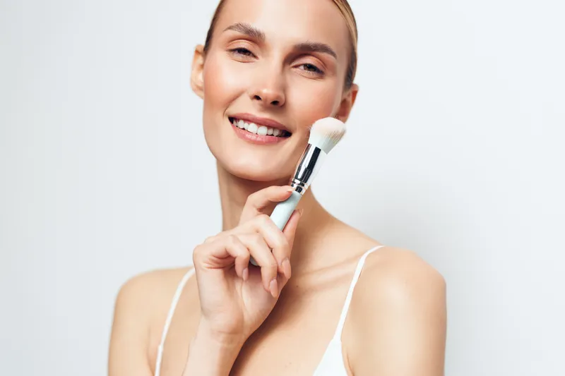 Smiling young woman with clear skin holds makeup brush near face, demonstrating natural beauty and skincare routine, isolated on white background with positive expression.