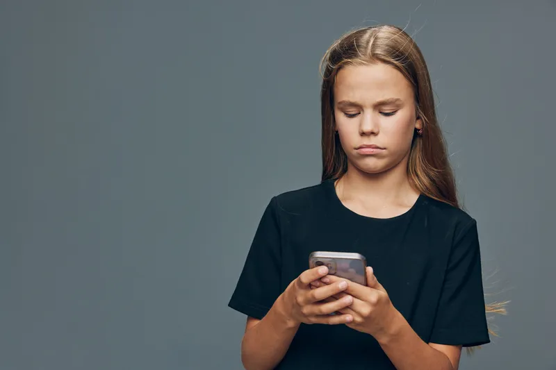 Girl with long hair wearing black shirt looking at smartphone screen with serious expression on gray background. Youth, technology and concentration with mobile device in hands.