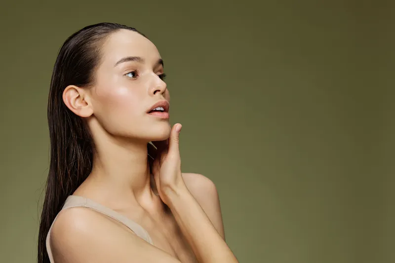 Elegant young woman with long hair touching her face on a green background, showcasing natural beauty and skincare