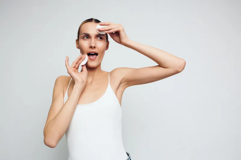 Young woman with clear skin removing makeup with cotton pads, natural beauty routine, white tank top, clean background, healthy skin care, fresh and minimalistic style