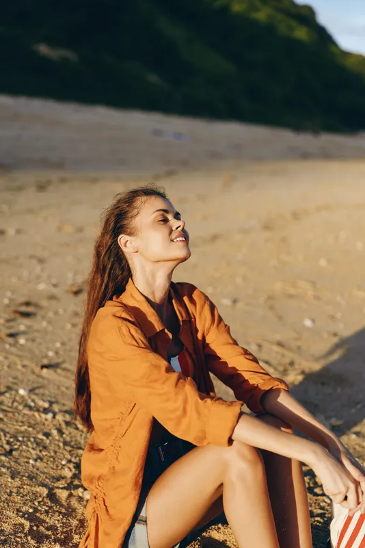 Young woman sits on a sunlit beach wearing an orange shirt, eyes closed, enjoying a warm breeze and a relaxed mood. Her hair falls loosely as the golden light highlights the sand and distant green