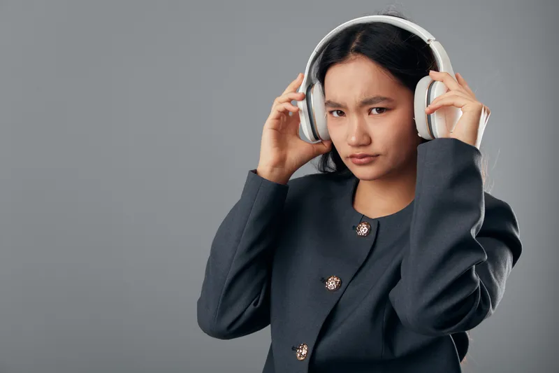 Teenage girl adjusting headphones with displeased expression on neutral background. Young female listening to music, wearing dark jacket with buttons for casual lifestyle and technology.