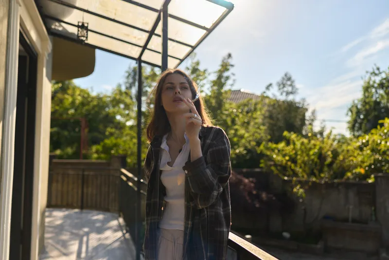 A woman in casual outdoor attire stands on a sunlit balcony, thoughtful pose, hand near chin, as she reflects in a gentle lifestyle moment, conveying introspection