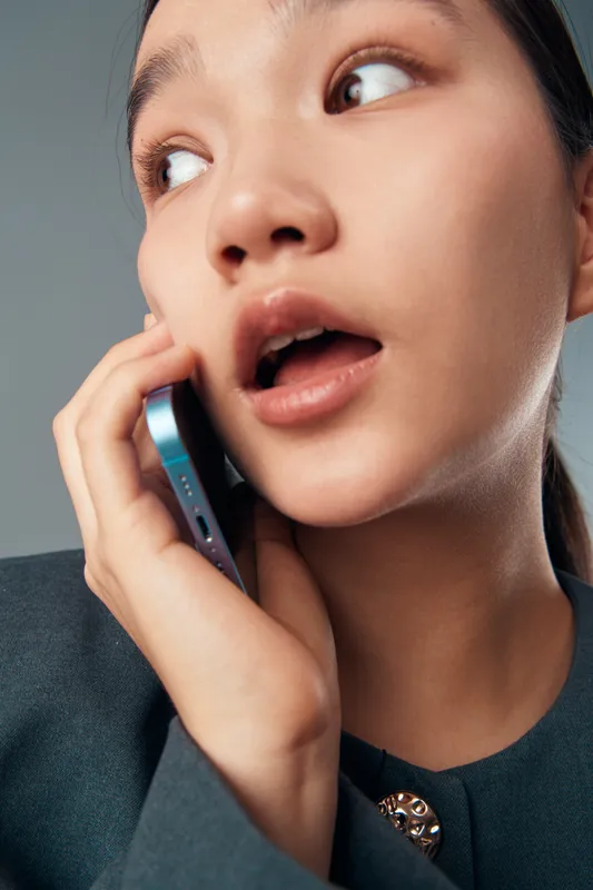 Young woman holding smartphone to ear with surprised expression in closeup portrait. Asian female talking on mobile phone with open mouth and wide eyes indoors with neutral background.