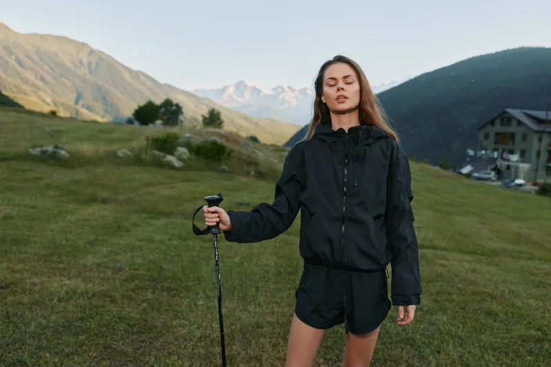 A confident young woman stands in a grassy field with a trekking pole, wearing a black windbreaker and shorts. She looks ahead, ready for a hike under clear skies.