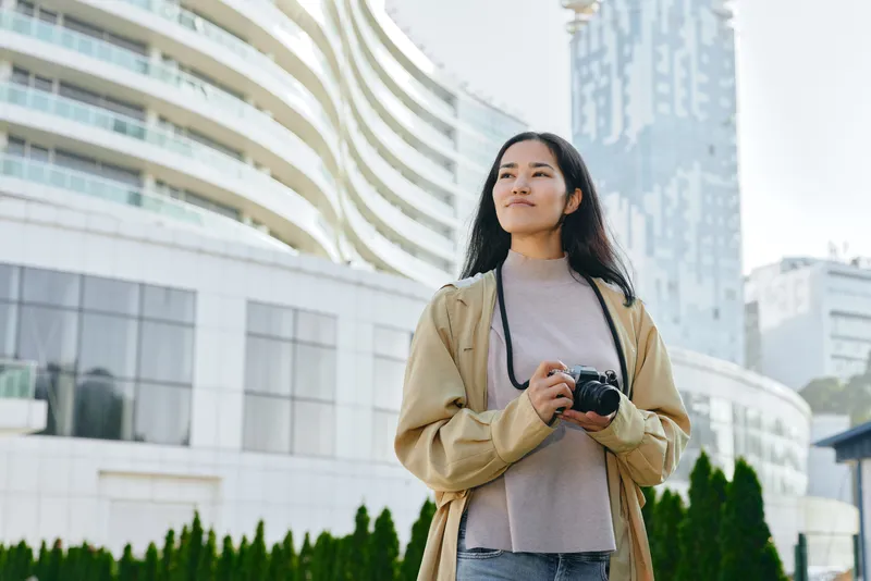 Woman with camera in urban streets, standing outdoors amid modern buildings, ready to shoot city landscapes, fashion style, and street photography in bright daylight ambience