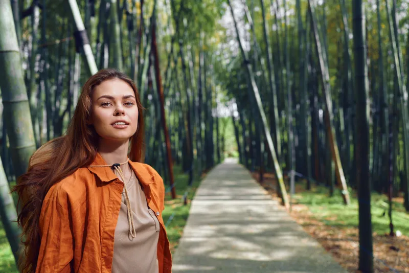 woman bamboo forest path portrait, traveler with backpack standing on paved walkway among tall bamboo stalks, wearing orange jacket in outdoor nature scene with calm, confident expression