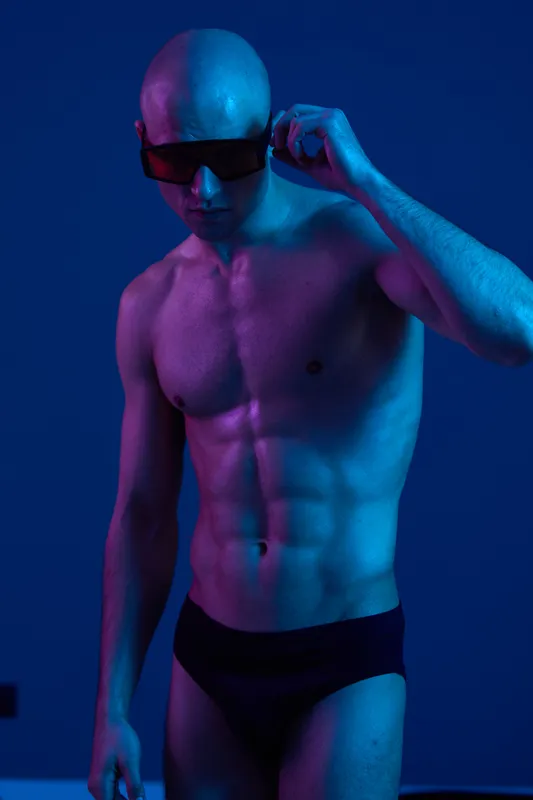 Athletic man wearing sunglasses poses against a vibrant blue and purple background, showcasing strength and confidence in an artistic light composition.