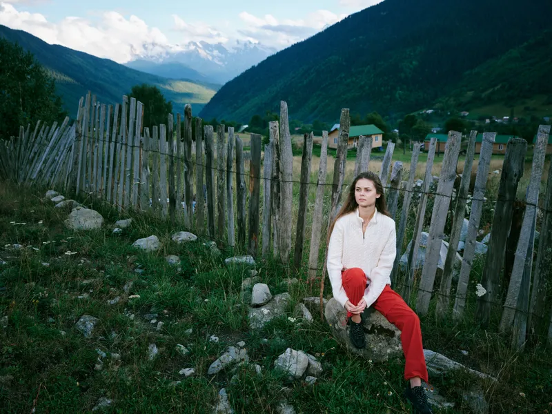 Woman in a white top sits beside a weathered wooden fence in a rural valley, red pants, mountain scenery, calm mood, natural light, peaceful countryside and distant peaks.