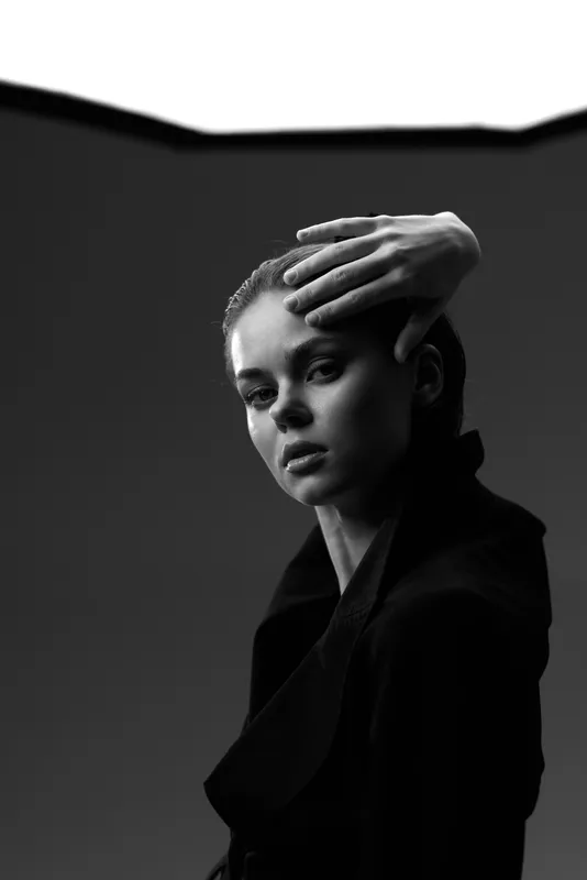 Black and white portrait of a serious woman with hand on forehead in dark clothing, dramatic lighting, and moody expression.