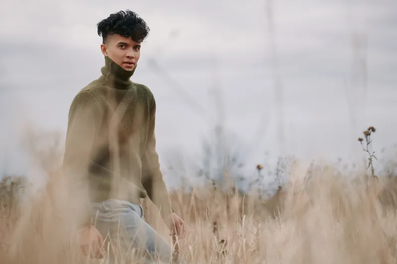 A solitary figure stands in tall grass, wearing a chunky knit turtleneck. The scene conveys authenticity through natural light, soft focus, and an unposed, honest mood.