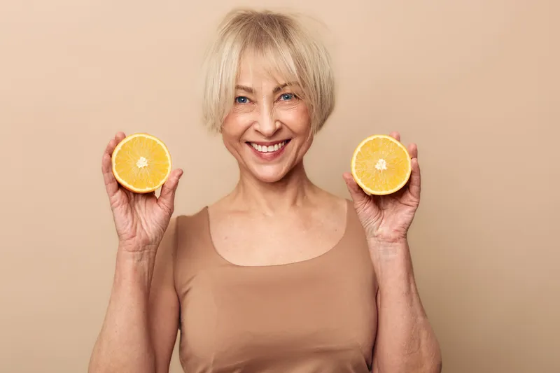 Smiling mature woman with short blonde hair holds two halves of fresh orange fruit, wearing beige tank top against neutral background. Concept for healthy lifestyle, natural vitamins, and wellness.
