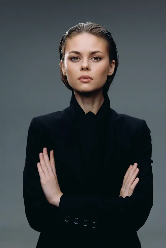 Serious woman in black jacket posing confidently with folded arms against gray background