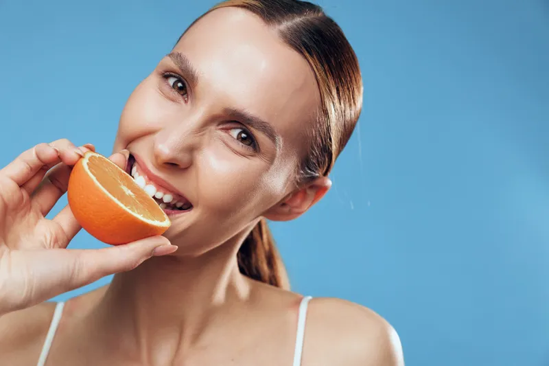 Woman orange citrus healthy smile closeup vitamin beauty concept, young woman in white straps drinks juice from orange, fresh face, blue studio background clean happy portrait.