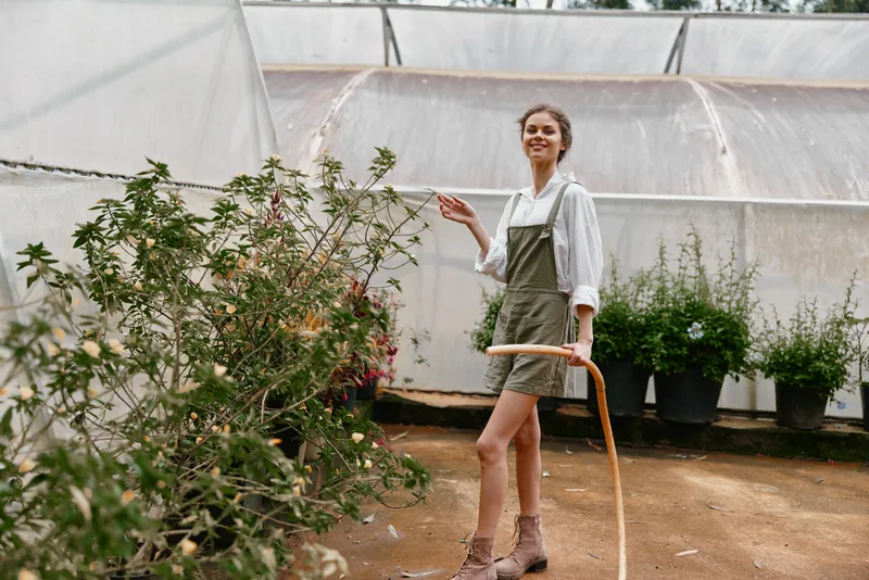 Young woman in casual overalls smiling while watering plants in a bright greenhouse, showcasing gardening joy and eco friendly lifestyle