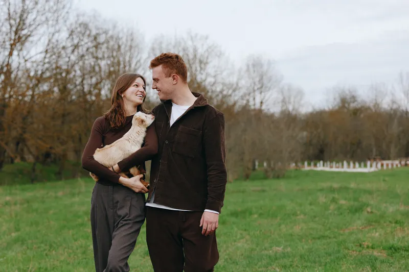 Joyful couple outdoors with a goat kid between them, casual attire, relaxed mood, countryside setting. Love, companionship, and warmth in smiles. Natural light and soft colors decorate the field.