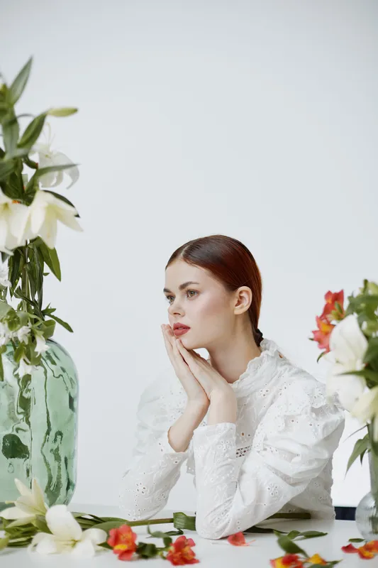 Elegant young woman with red hair in a white blouse, surrounded by fresh flowers in a minimalist setting, expressing contemplation and beauty
