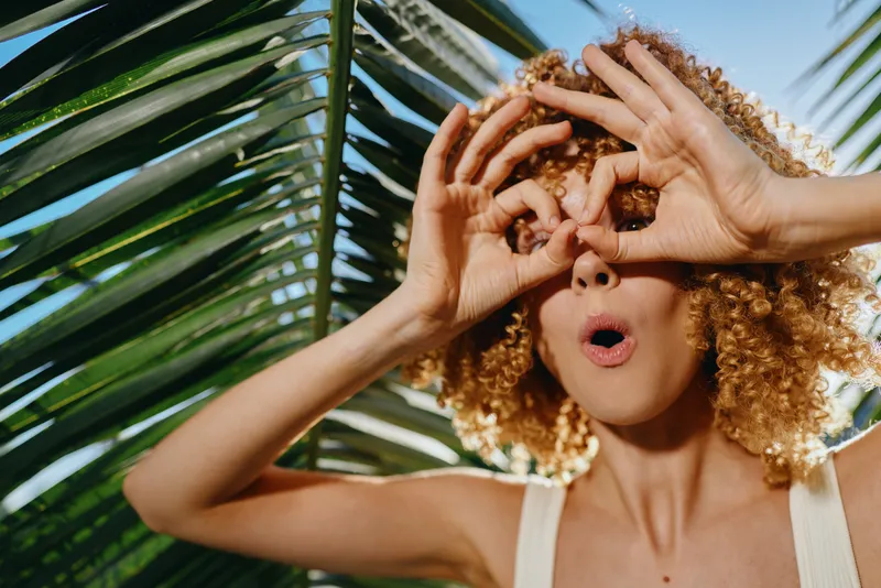 Young woman with curly hair making binocular gesture with hands near tropical palm leaves in bright sunlight, playful mood, summer outdoor fun and natural background.