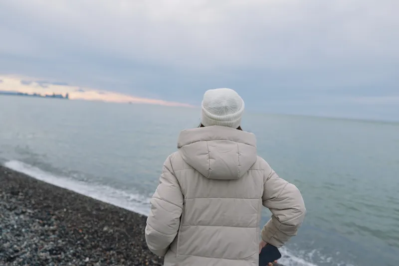 Person wearing a light winter coat gazes at the sea along a pebbled coastline, the calm water stretching to the horizon under a soft cloudy sky, capturing solitude and contemplation by the shore.
