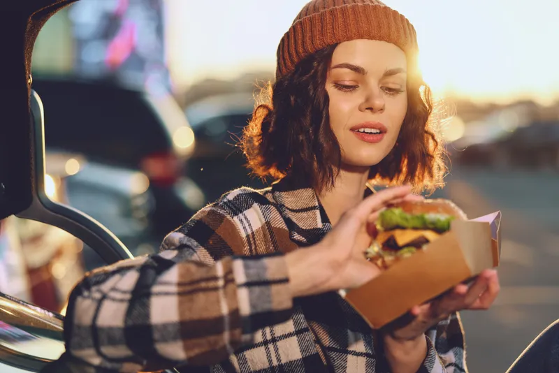 Woman with burger by car at golden hour, beanie and candid outdoor portrait conveying authenticity, candid lifestyle, golden hour glow, mindful living and emotional storytelling.