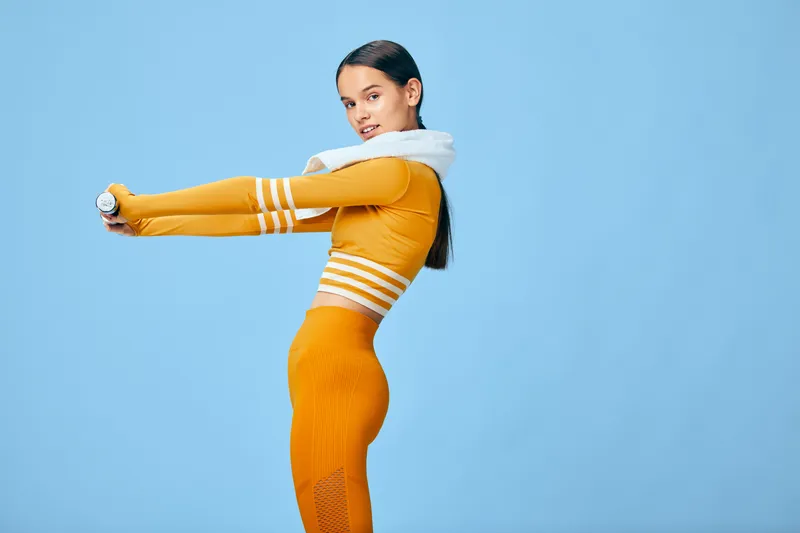 Woman in orange fitness outfit performing a forward stretch during a workout, showcasing athleticism, balance and concentration against a vibrant blue backdrop.
