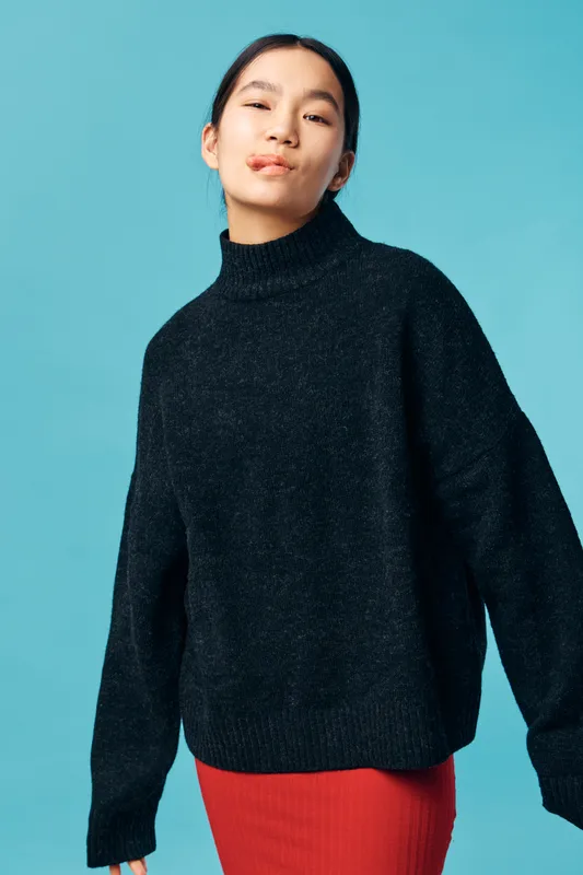 Young woman wearing black turtleneck sweater and red skirt posing confidently with slight smile against blue background. Female model stands with relaxed posture and calm expression in studio.