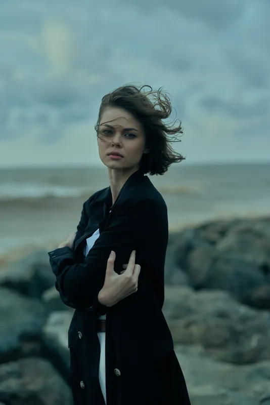 Moody coastal scene with wind sweeping through a woman wearing a dark jacket along a rocky shoreline, ocean in the background, clouds overhead, creating a tense atmospheric moment.