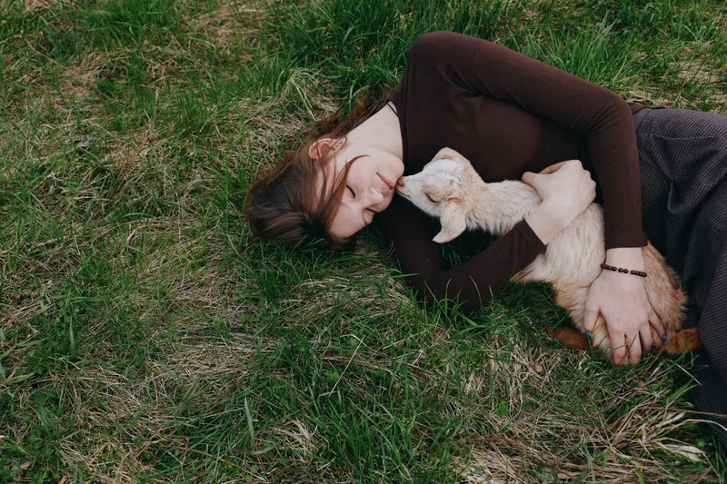 Relaxed woman lying on grass, cuddling a small goat kid under soft daylight. Rural lifestyle moment, gentle touch, connection with nature, calm warmth and wellbeing. The scene conveys comfort,
