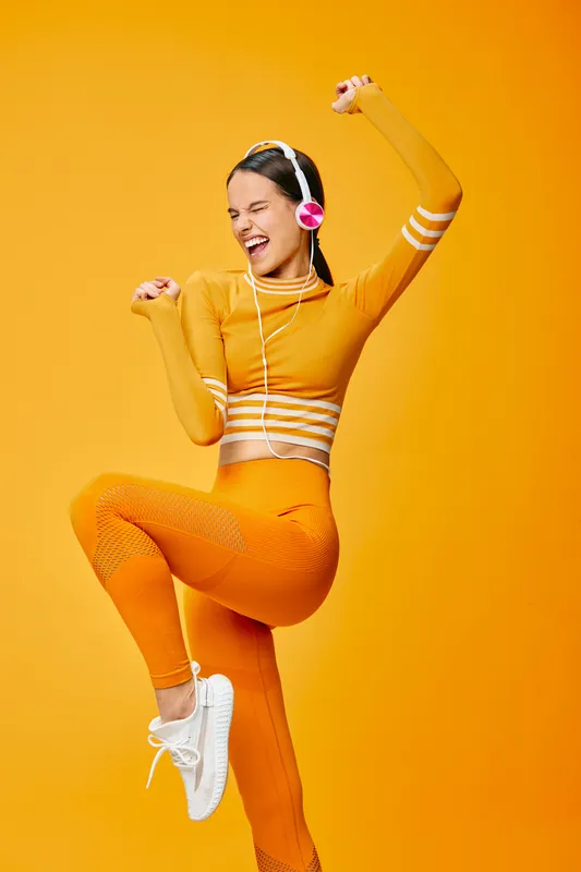 Energetic woman in bright orange athletic wear dances with headphones, expressing fitness and vitality in a bold studio setting. Her smile and dynamic pose convey confidence, movement, and wellness