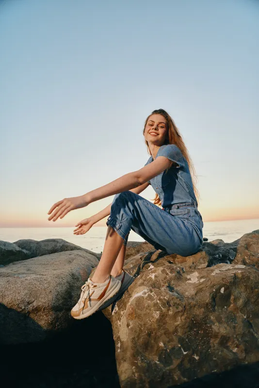 smiling woman sitting on rocks by the sea during sunset wearing casual denim outfit and sneakers, relaxed outdoor atmosphere with clear sky and warm light