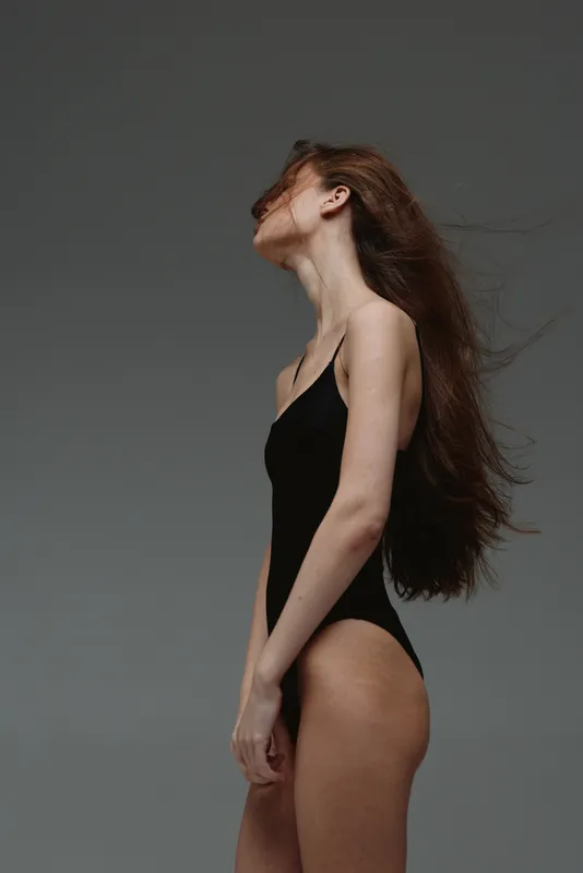 Woman posing in black swimsuit with long flowing hair against a dark gray studio background, showcasing elegance and natural beauty.