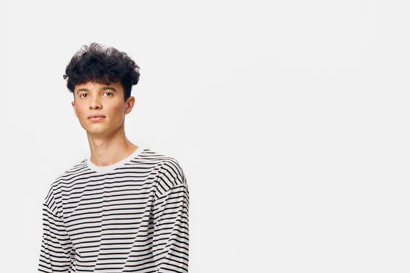 young man with curly hair wearing striped shirt stands against white background, casual pose, natural expression, trendy hairstyle with soft lighting