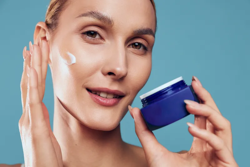 Young woman applies moisturizing cream on her face holding blue jar close to skin with light makeup and natural expression isolated on blue background.