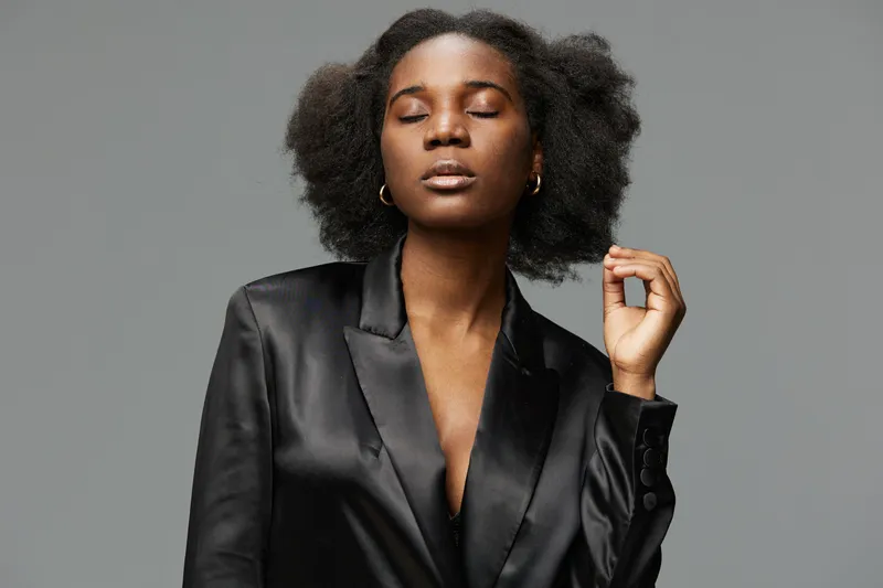 Elegant woman with natural afro hairstyle in a black satin blazer, expressing confidence against a minimal gray background