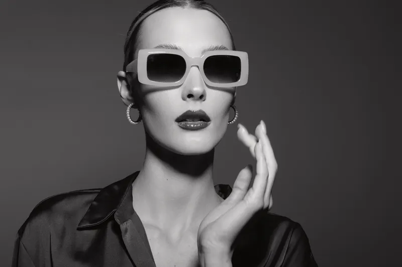 Fashionable caucasian woman with pale skin wearing large sunglasses and earrings poses in black shirt on dark background studio portrait.