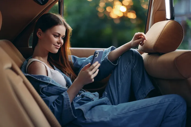 Young woman in casual denim outfit enjoying her smartphone in a cozy car setting, capturing a relaxed mood during sunset.