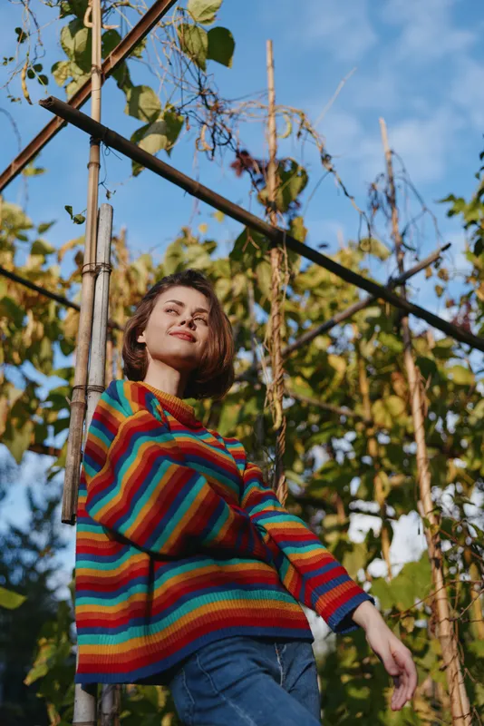 Young woman in her mid 20s with short hair, smiling and leaning on a wooden trellis in a sunlit garden. Casual outfit with colorful striped sweater and jeans, relaxed lifestyle portrait.