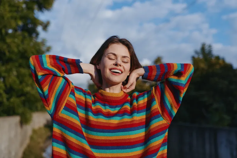 Young woman in a colorful striped sweater smiling with eyes closed outdoors, relaxed pose with hands behind head, casual outfit, sunny blue sky and joyful lifestyle vibe.